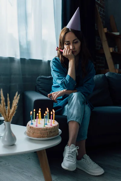 Сумна жінка тримає партійний ріг, сидить на дивані і дивиться на торт на день народження на кавовому столі — стокове фото