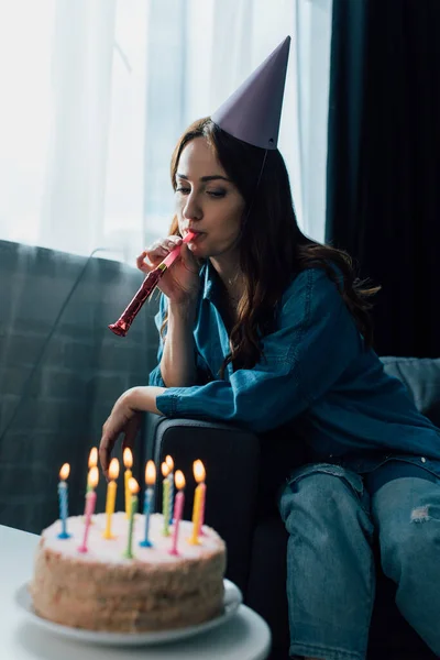 Селективное внимание грустной женщины, дующей в рог вечеринки и сидящей на диване рядом с праздничным тортом на журнальном столике — стоковое фото