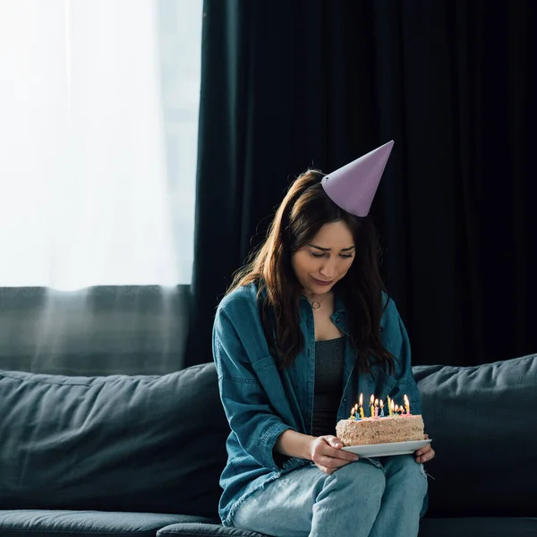 Frustrated woman in party cap sitting on sofa and holding birthday cake with candles — Stock Photo