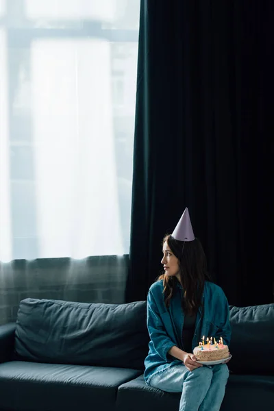 Засмучена жінка у вечірній кепці, сидячи на дивані, тримає торт на день народження зі свічками і дивиться в сторону — стокове фото