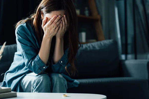 Mujer molesta cubriendo la cara mientras está sentado cerca de la mesa de café con anillo de oro, concepto de divorcio - foto de stock