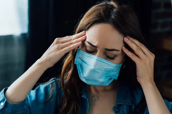 Втомлена брюнетка жінка в медичній масці торкається голови вдома — стокове фото