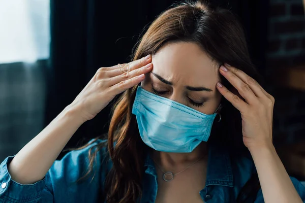 Femme brune fatiguée en masque médical touchant la tête à la maison — Photo de stock