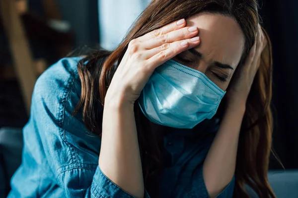 Brunette woman in medical mask touching head while having migraine at home — Stock Photo