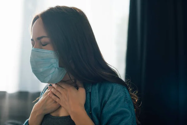 Morena mujer con los ojos cerrados en máscara médica tocando garganta en casa - foto de stock