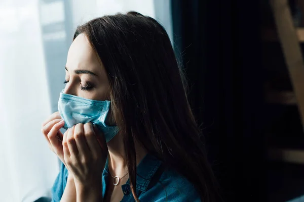 Mulher morena com olhos fechados tocando máscara médica em casa — Fotografia de Stock