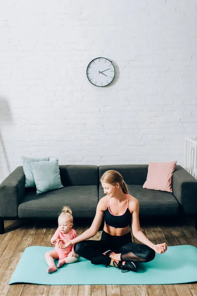 Madre en ropa deportiva sentada en postura de yoga cerca de la hija lactante en la esterilla de fitness - foto de stock