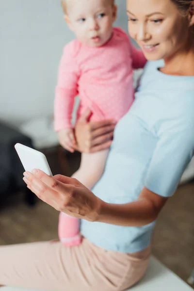 Foco seletivo da mulher usando celular e segurando o bebê em casa — Fotografia de Stock