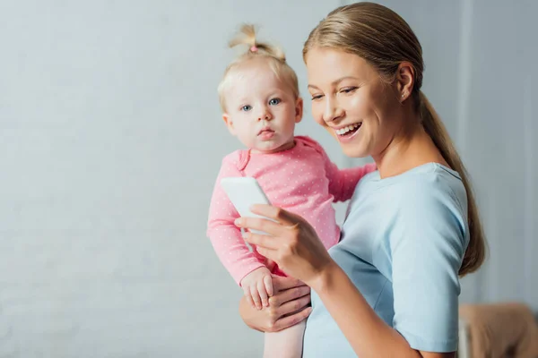 Selective focus of woman using smartphone while baby daughter looking at camera — Stock Photo