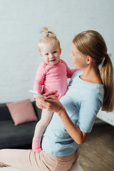 Concentration sélective de la femme tenant smartphone et bébé fille à la maison — Photo de stock