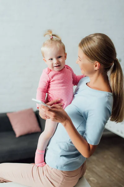 Enfoque selectivo de la mujer sosteniendo el teléfono inteligente y la niña en casa - foto de stock