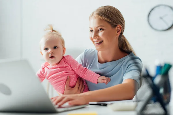 Foco seletivo da mãe segurando bebê menina e usando laptop em casa — Fotografia de Stock