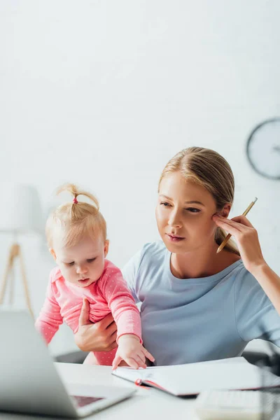 Selective focus of concentrated woman holding pencil and infant while working with laptop at table — Stock Photo