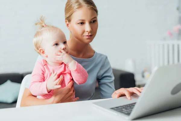 Selective focus of woman holding daughter and using laptop at table — Stock Photo