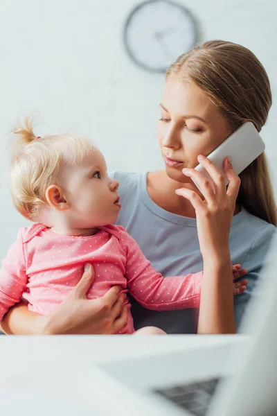Foco seletivo da mulher falando no smartphone e olhando para a filha infantil perto do laptop na mesa — Fotografia de Stock