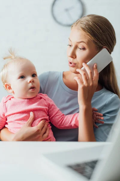 Enfoque selectivo de la madre sosteniendo a la niña y hablando en el teléfono inteligente mientras trabaja en casa - foto de stock