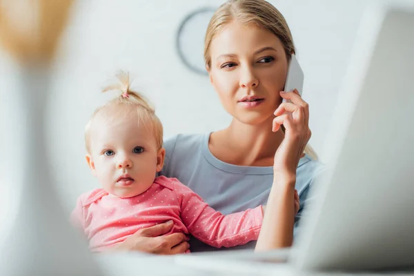 Selective focus of focused woman talking on smartphone and holding daughter at home — Stock Photo