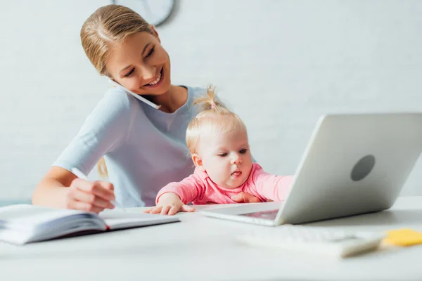 Selective focus of infant sitting near mother talking on smartphone and working at home — Stock Photo