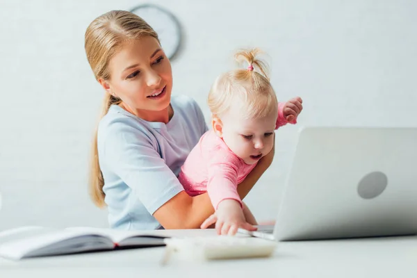 Selective focus of woman holding infant daughter while working with notebook and laptop at home — Stock Photo