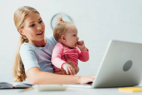 Enfoque selectivo de la madre sosteniendo a la niña mientras trabaja en la computadora portátil en casa - foto de stock