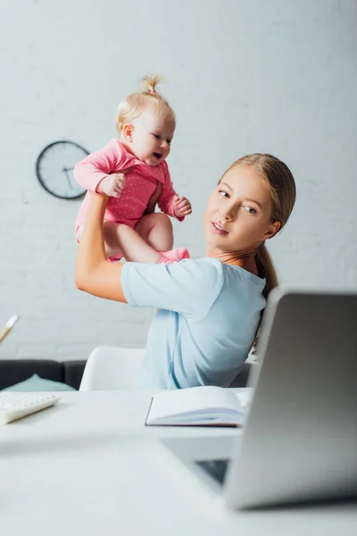 Selective focus of woman playing with baby girl while looking at laptop on table at home — Stock Photo