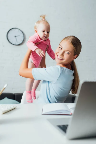 Enfoque selectivo de la mujer mirando a la computadora portátil mientras sostiene a la hija infantil en la mesa - foto de stock