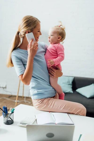 Focus selettivo della madre che guarda la bambina mentre parla sullo smartphone vicino alla cancelleria e al laptop sul tavolo — Foto stock