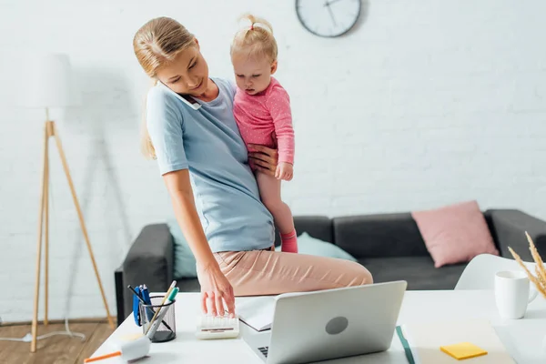 Selective focus of woman talking on smartphone and using calculator while holding daughter at home — Stock Photo