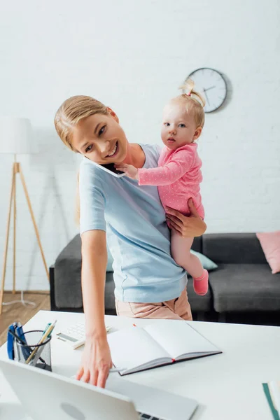 Selective focus of woman holding baby girl while talking on smartphone and using laptop at home — Stock Photo
