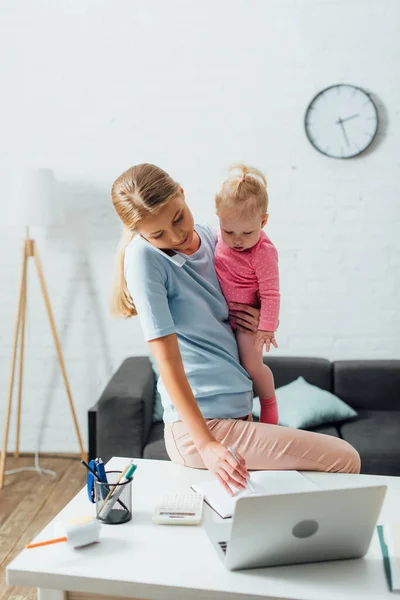 Selective focus of freelancer talking on smartphone and holding infant daughter while working at home — Stock Photo