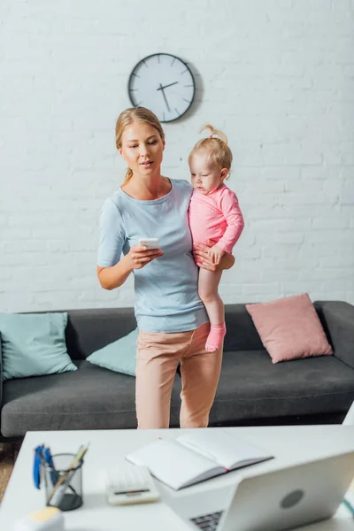 Selective focus of woman holding smartphone and baby girl near stationery and laptop on table — Stock Photo