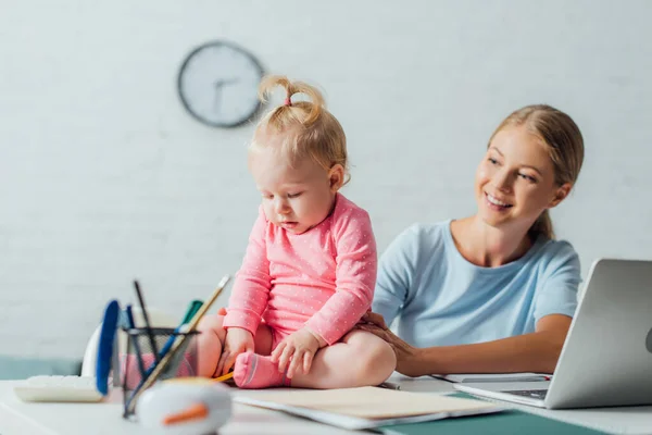 Selective focus of baby girl sitting near stationery and laptop on table beside mother — Stock Photo