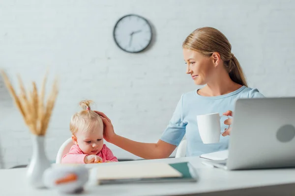 Selective focus of freelancer with cup touching infant daughter near laptop and stationery on table — Stock Photo