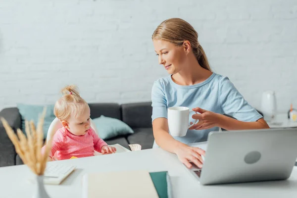 Enfoque selectivo de la mujer sosteniendo la taza y usando el ordenador portátil cerca de la niña en casa - foto de stock