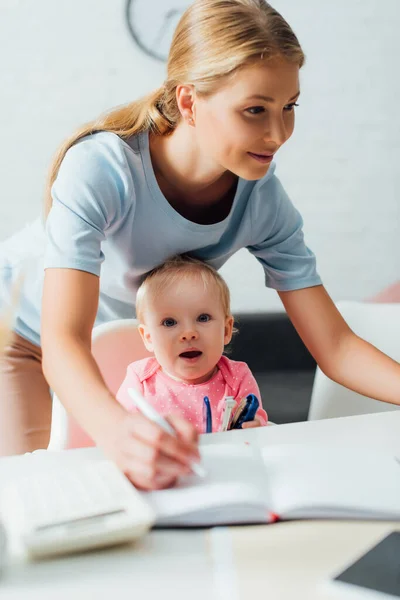 Focus selettivo di bambino tenendo cucitrice mentre la madre scrive sul taccuino a casa — Foto stock