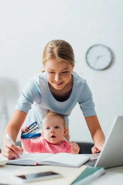 Concentration sélective de la mère travaillant avec un ordinateur portable et écrivant sur un ordinateur portable pendant que l'enfant tient une agrafeuse à table — Photo de stock