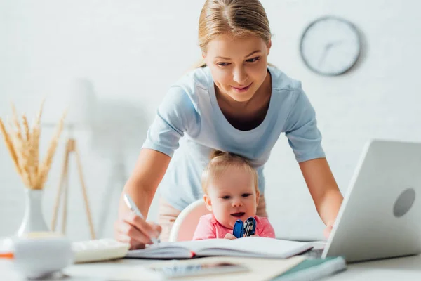 Enfoque selectivo de la mujer que escribe en el cuaderno mientras trabaja cerca del bebé con grapadora en la mesa - foto de stock