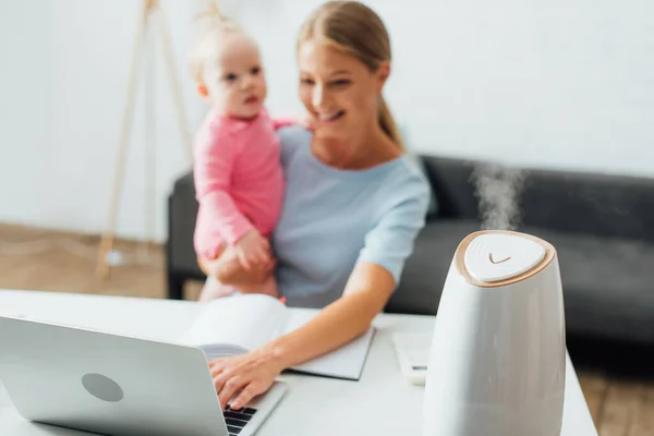 Foco seletivo de umidificador perto da mulher usando laptop e segurando a filha do bebê — Fotografia de Stock