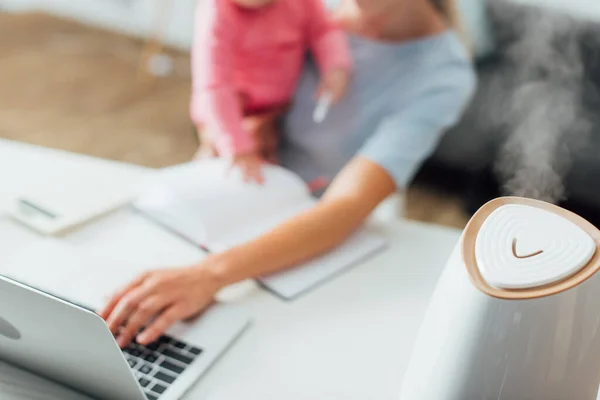 Enfoque selectivo del humidificador en la mesa cerca de la mujer que trabaja con el ordenador portátil y sostiene a la niña en segundo plano - foto de stock