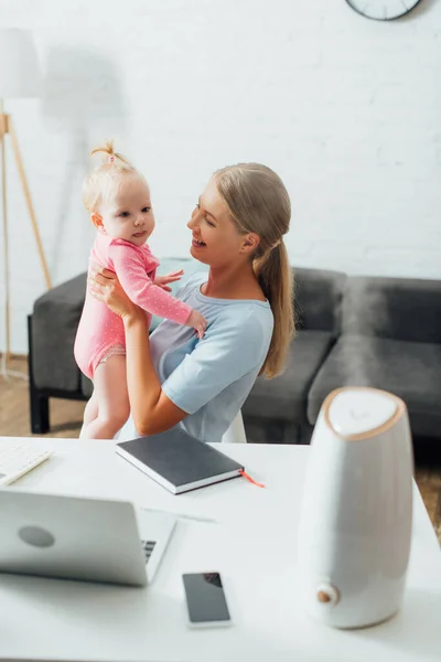 Selective focus of mother holding baby girl near gadgets, notebook and humidifier on table — Stock Photo