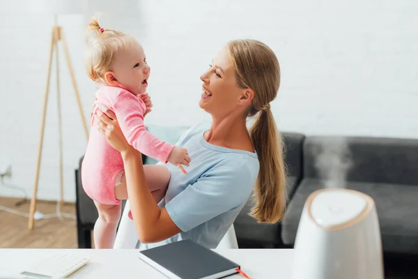 Focus selettivo della donna che tiene il bambino vicino al notebook e all'umidificatore sul tavolo — Foto stock