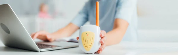 Panoramic crop of woman holding baby monitor while using laptop at table — Stock Photo