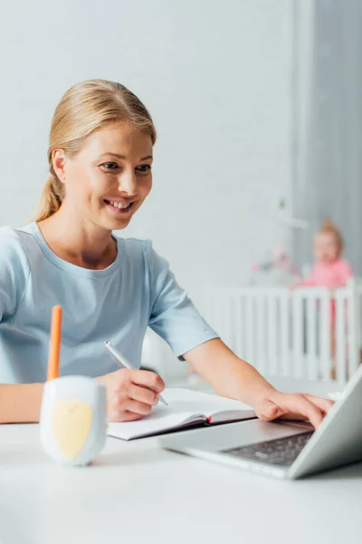 Selective focus of woman using laptop and writing on notebook near baby monitor on table — Stock Photo