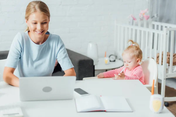 Foco seletivo de bebê menina segurando caneta enquanto a mãe usando laptop em casa — Fotografia de Stock