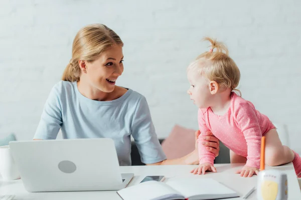 Selektiver Fokus der Frau, die ihre Tochter ansieht, während sie mit Laptop in der Nähe des Babyfons auf dem Tisch arbeitet — Stockfoto