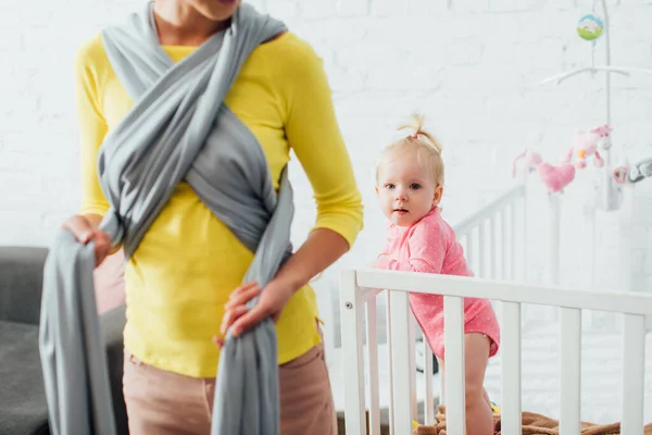 Selective focus of infant girl in baby crib near mother holding sling at home — Stock Photo