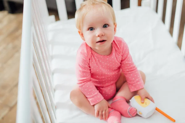 Selective focus of infant girl holding baby monitor while sitting in crib — Stock Photo