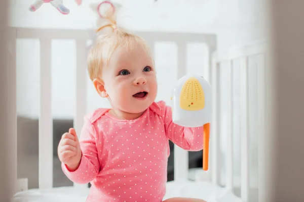 Selective focus of excited infant girl with baby monitor looking away in crib — Stock Photo