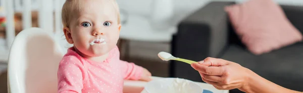 Panoramic crop of woman holding spoon with baby food near daughter with messy mouth on feeding chair — Stock Photo