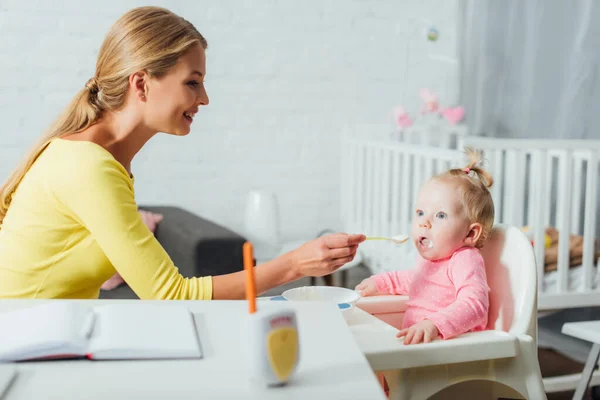 Foco seletivo da mãe alimentando bebê menina na cadeira alta perto de notebook na mesa — Fotografia de Stock
